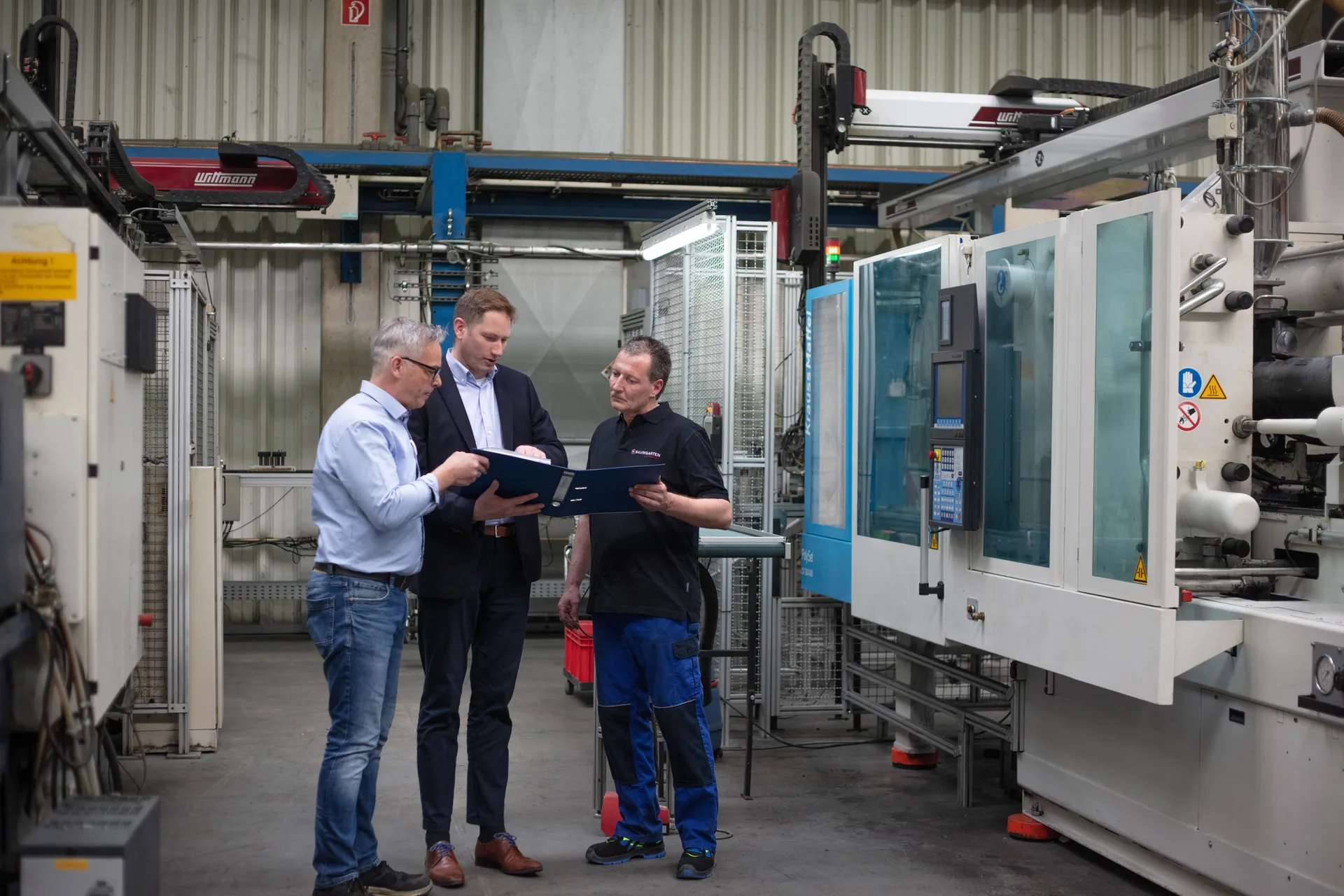 Drei Mitarbeiter besprechen ein Projekt mit Unterlagen vor einer Spritzgießmaschine